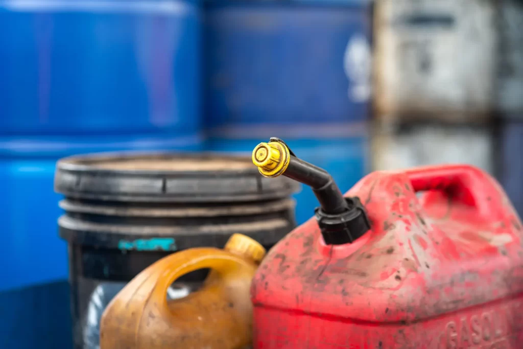 tank2 - Techsvět Detailní záběr na červený plastový kanystr s nálevkou určený na převoz benzínu nebo nafty. V pozadí jsou vidět další nádoby a barely.