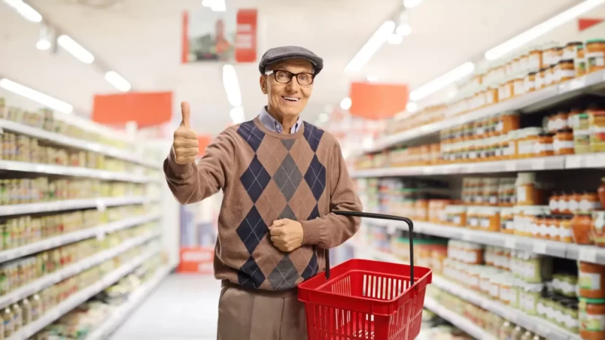 Usměvavý senior v supermarketu, který drží červený nákupní košík a ukazuje palec nahoru.