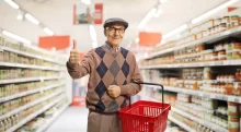 Usměvavý senior v supermarketu, který drží červený nákupní košík a ukazuje palec nahoru.