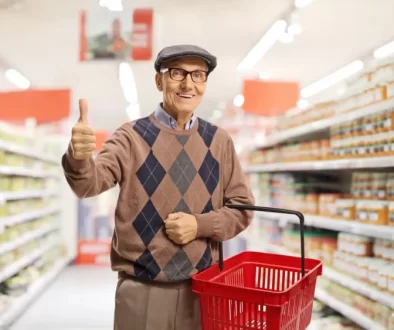 Usměvavý senior v supermarketu, který drží červený nákupní košík a ukazuje palec nahoru.