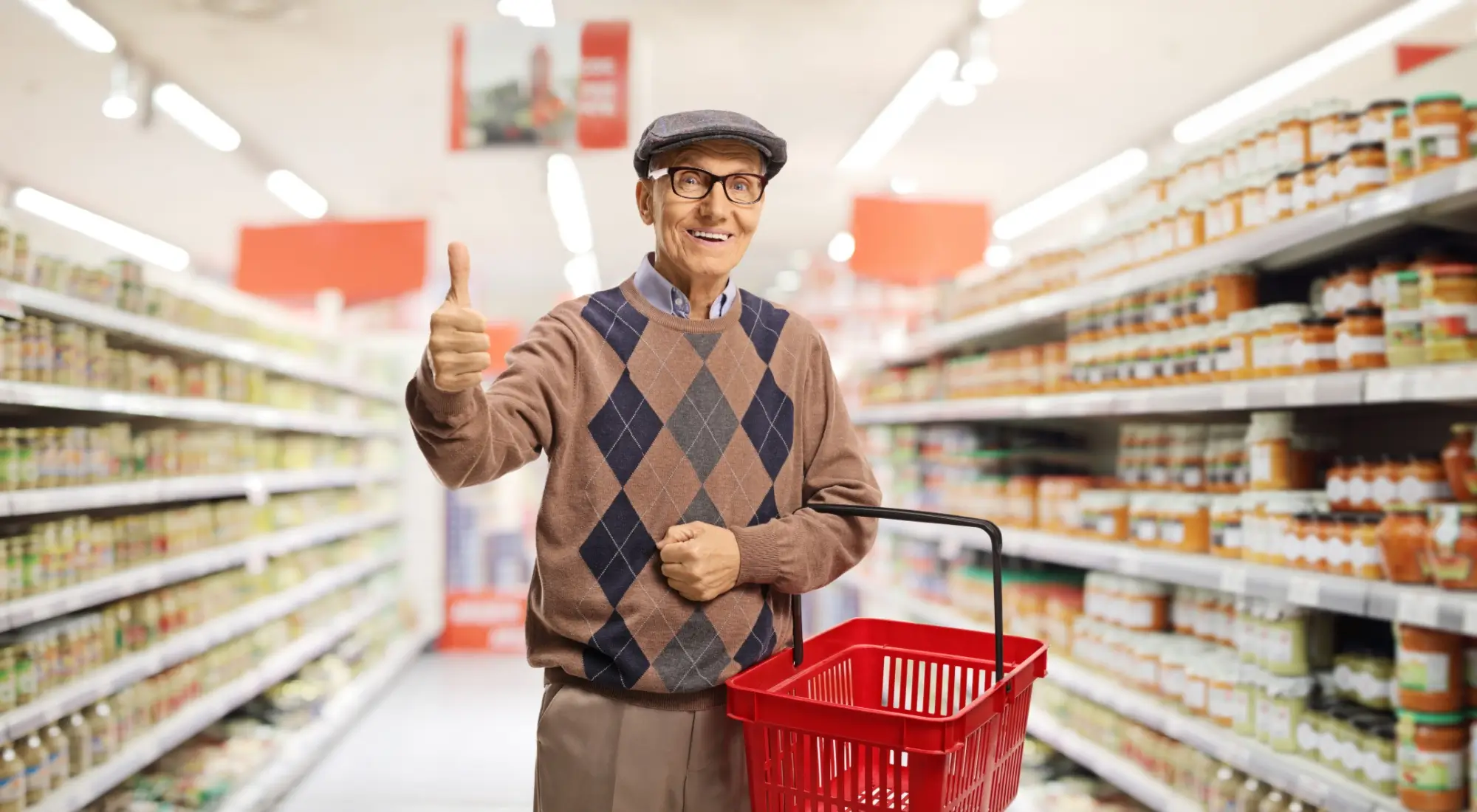 Usměvavý senior v supermarketu, který drží červený nákupní košík a ukazuje palec nahoru.