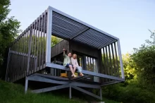 Moderní tiny house postavený na dřevěné konstrukci ve svahu, obklopený přírodou.
