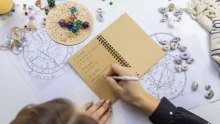 Pracovní stůl, kde žena počítá numerologické hodnoty do zápisníku. Kolem jsou rozloženy astrologické diagramy, kameny s čísly, barevné krystaly, tarotové pomůcky a zapálená svíčka.