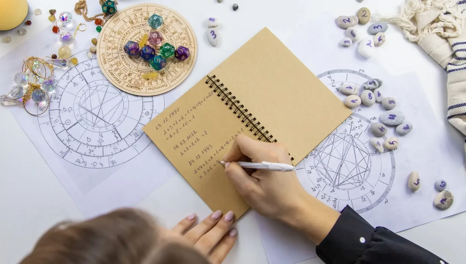 Pracovní stůl, kde žena počítá numerologické hodnoty do zápisníku. Kolem jsou rozloženy astrologické diagramy, kameny s čísly, barevné krystaly, tarotové pomůcky a zapálená svíčka.