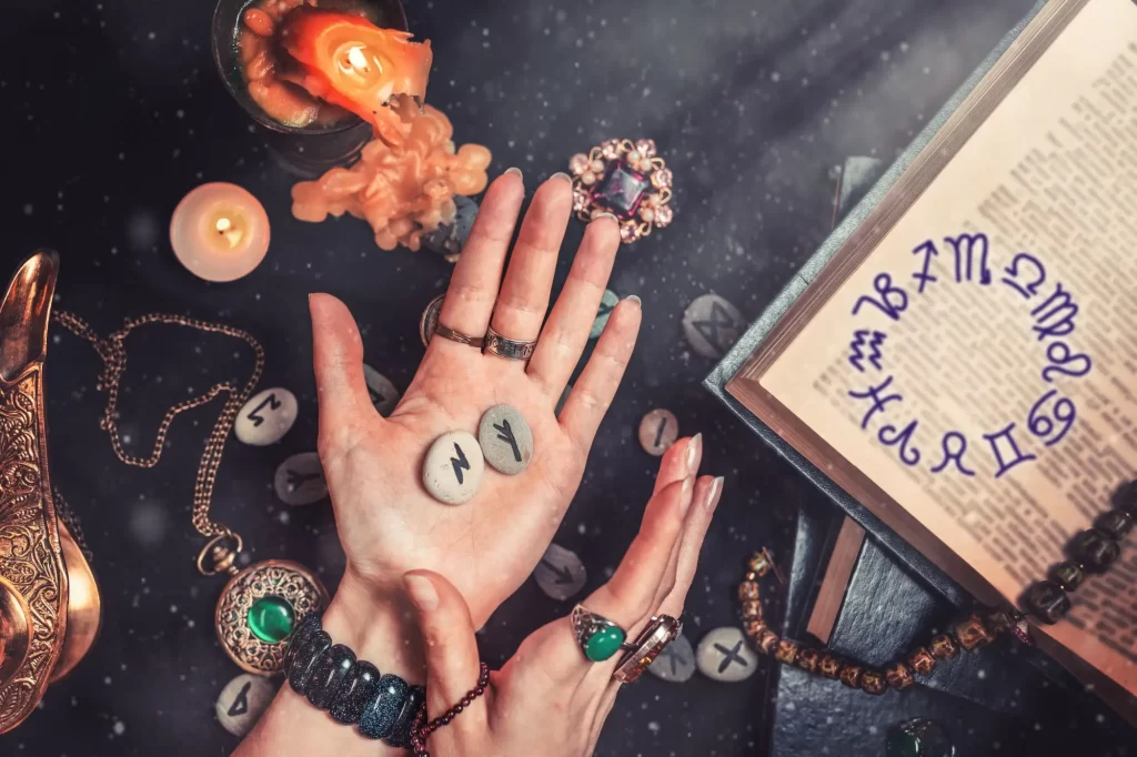 vědma3 - Techsvět Ruka věštkyně, která drží dva runové kameny, obklopená mystickými předměty jako svíčky, šperky, staré knihy a astrologické symboly.