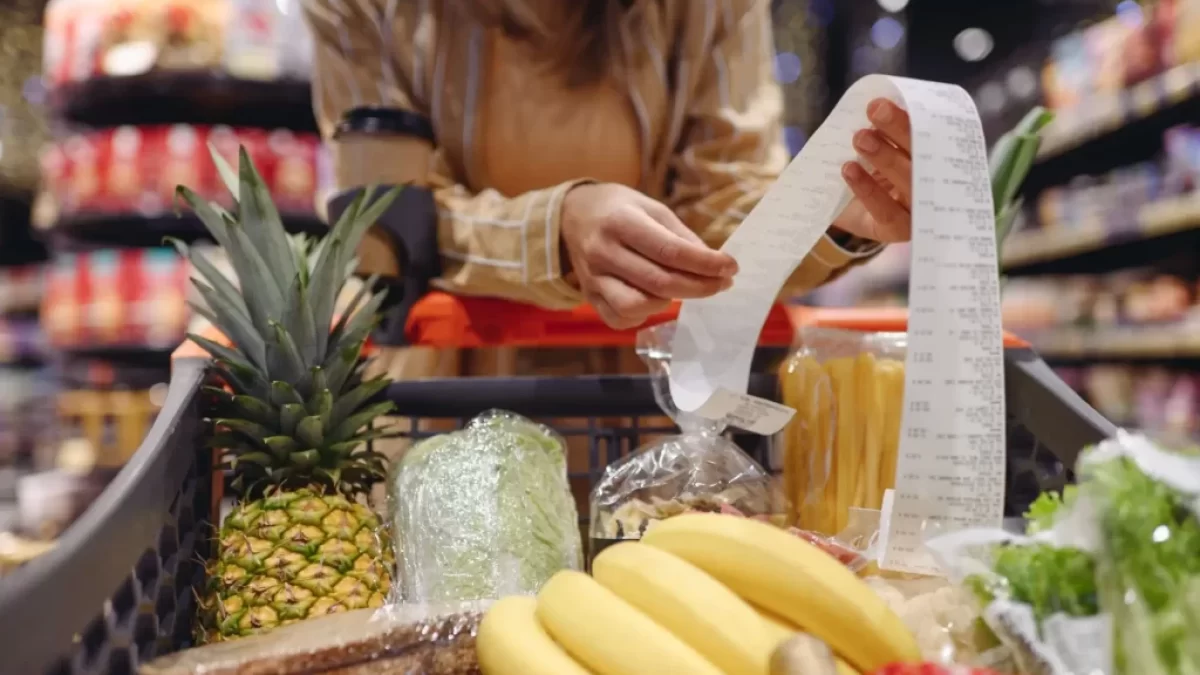 Žena v supermarketu, kontroluje dlouhý nákupní účet u plného vozíku s potravinami.