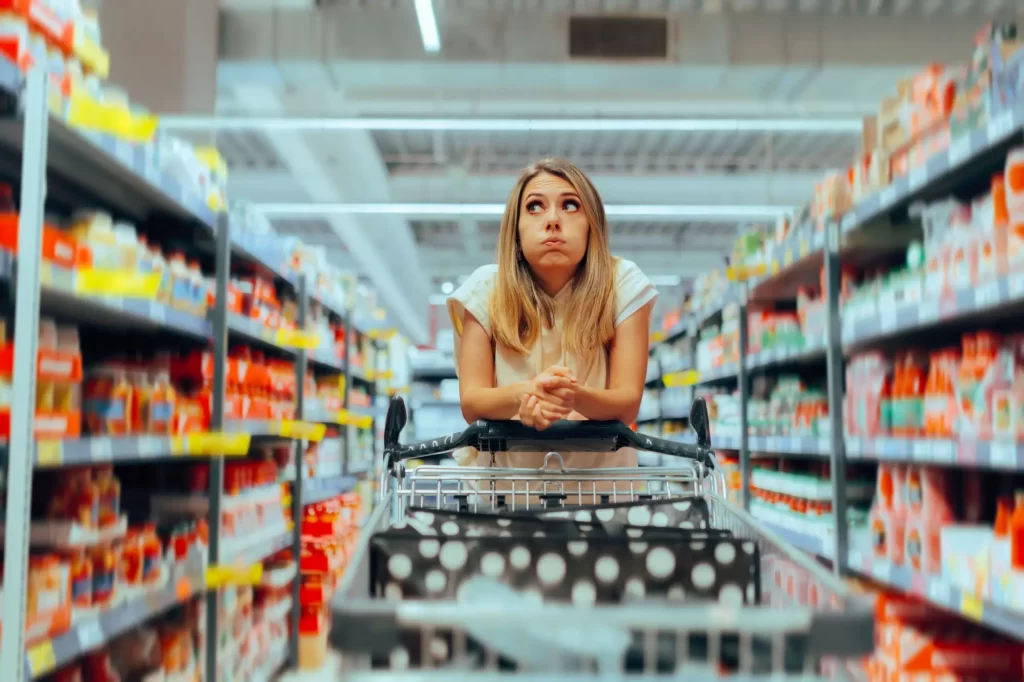 zboží3 - Techsvět Žena opřená o nákupní vozík uprostřed supermarketové uličky, s výrazem unavení nebo rozčarování.