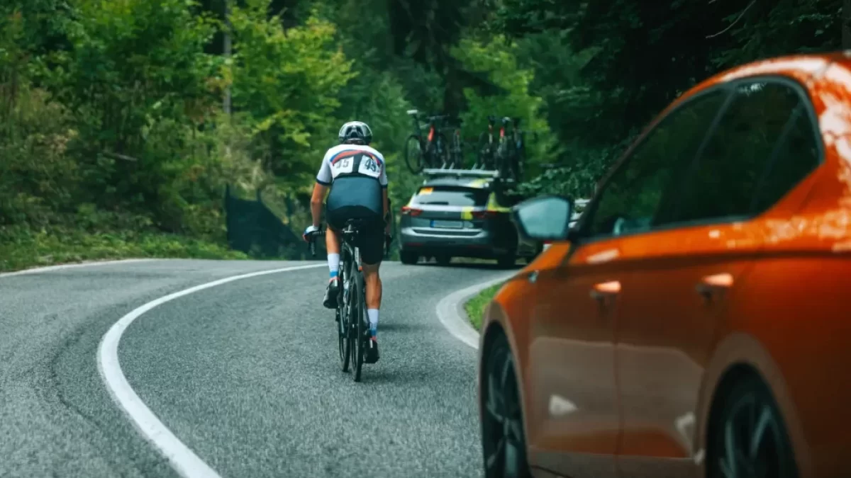 Dopravní situace na klikaté silnici v lese, kde jede cyklista vpravo po krajnici, před ním další vozidlo s koly na střeše. V levém dolním rohu je vidět část oranžového osobního auta, které se k cyklistovi blíží zezadu.