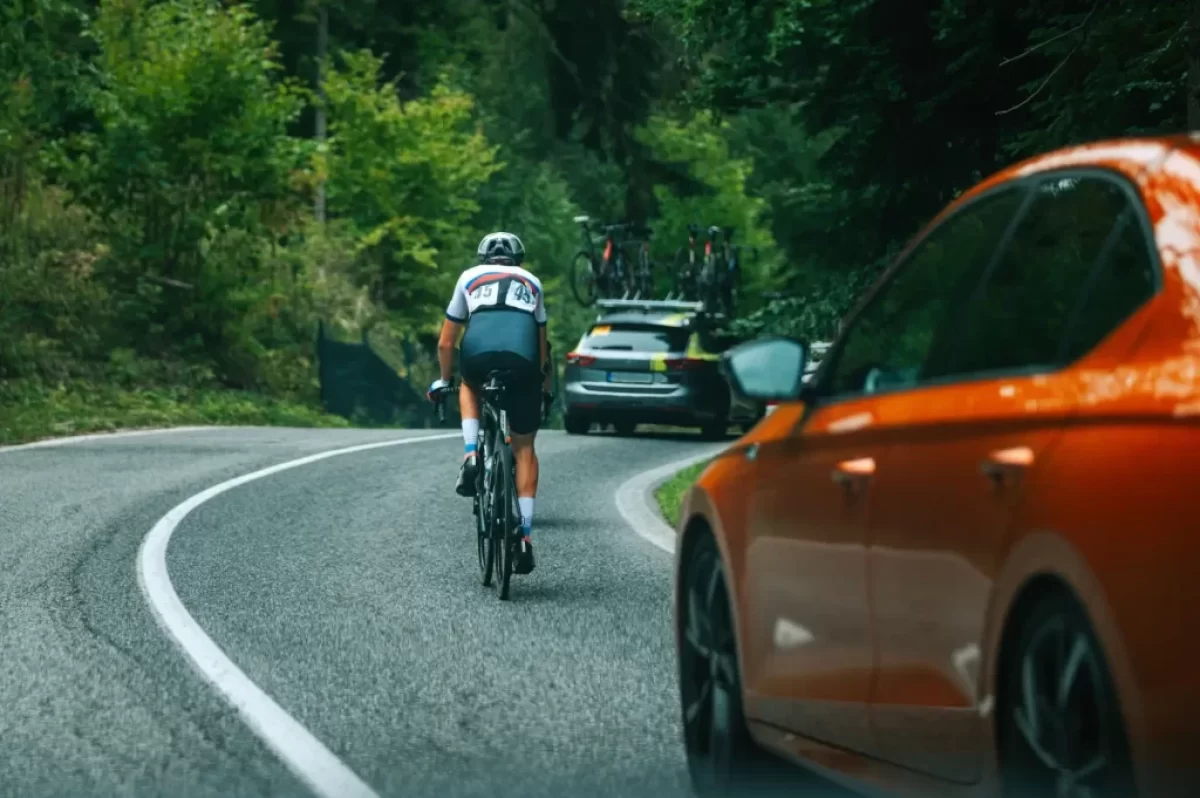 Dopravní situace na klikaté silnici v lese, kde jede cyklista vpravo po krajnici, před ním další vozidlo s koly na střeše. V levém dolním rohu je vidět část oranžového osobního auta, které se k cyklistovi blíží zezadu.