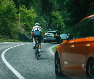 Dopravní situace na klikaté silnici v lese, kde jede cyklista vpravo po krajnici, před ním další vozidlo s koly na střeše. V levém dolním rohu je vidět část oranžového osobního auta, které se k cyklistovi blíží zezadu.