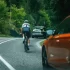Dopravní situace na klikaté silnici v lese, kde jede cyklista vpravo po krajnici, před ním další vozidlo s koly na střeše. V levém dolním rohu je vidět část oranžového osobního auta, které se k cyklistovi blíží zezadu.
