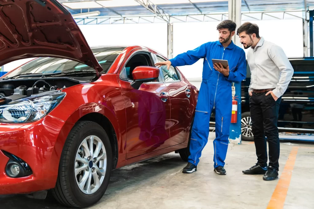 STK2 - Techsvět Automechanik v modré kombinéze, který pomocí tabletu ukazuje majiteli vozu určité technické detaily na červeném autě s otevřenou kapotou.