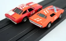 Dvě modelová auta na autodráze. Obě mají sportovní vzhled a jsou nalakovaná v oranžovo-červených barvách.