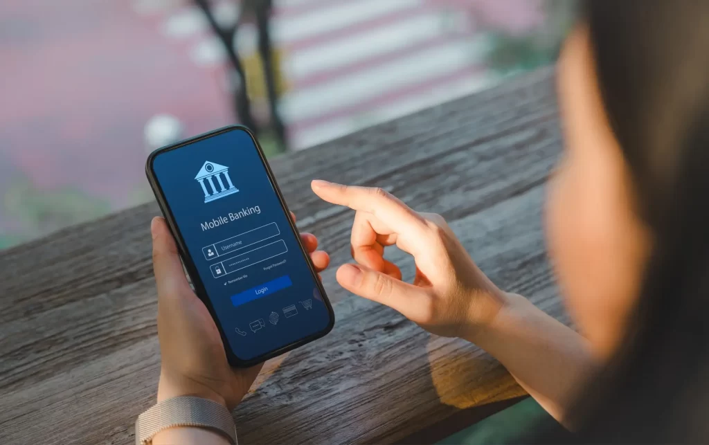 banka1 - Techsvět Žena se právě přihlašuje do mobilního bankovnictví na chytrém telefonu. Na displeji je zobrazené přihlašovací rozhraní s místem pro uživatelské jméno a heslo.