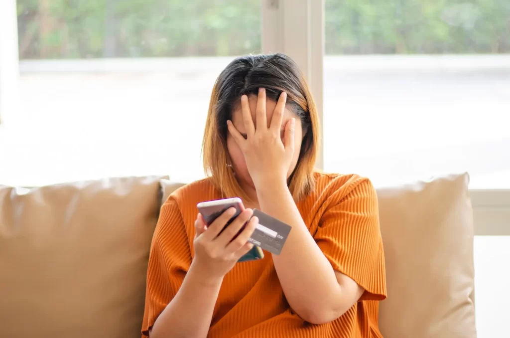 banka2 - Techsvět Žena sedící na pohovce, která si jednou rukou drží mobilní telefon a platební kartu a druhou si zakrývá obličej.