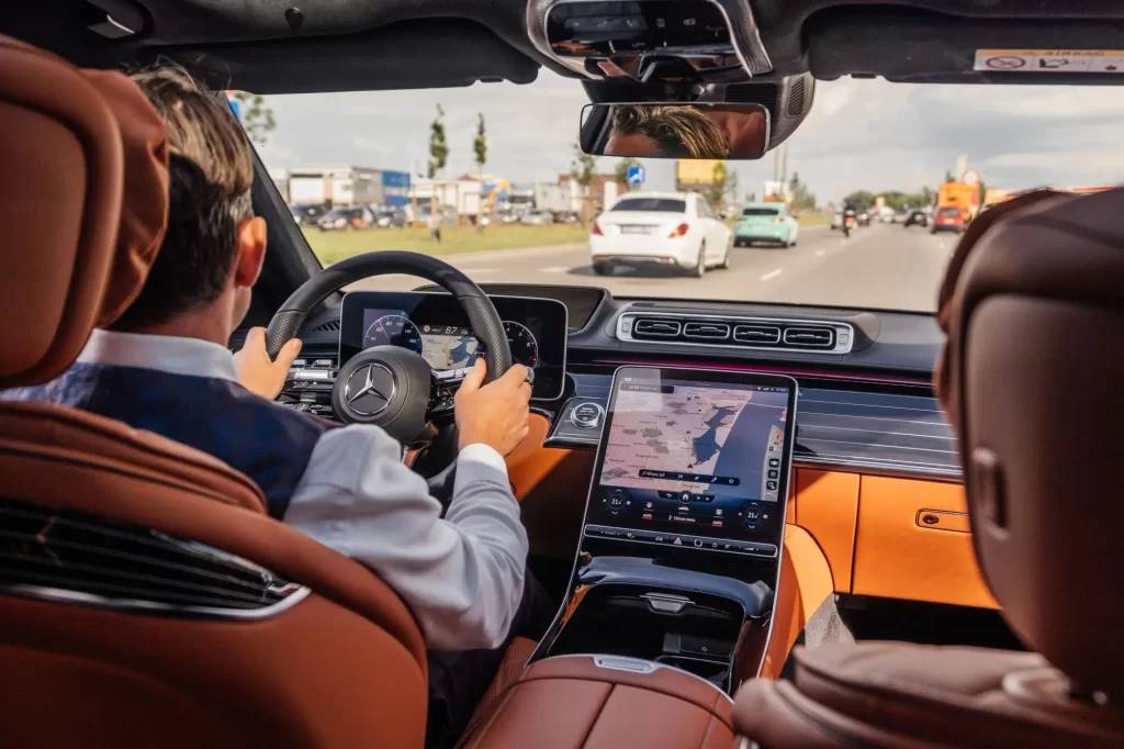 car3 - Techsvět Pohled ze zadního sedadla luxusního vozu Mercedes. Řidič v elegantní košili a vestě pevně drží volant, zatímco na středové konzoli svítí navigace.