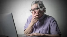 Starší muž v brýlích sedí u stolu a dívá se zamyšleně na otevřený notebook.