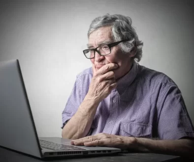 Starší muž v brýlích sedí u stolu a dívá se zamyšleně na otevřený notebook.