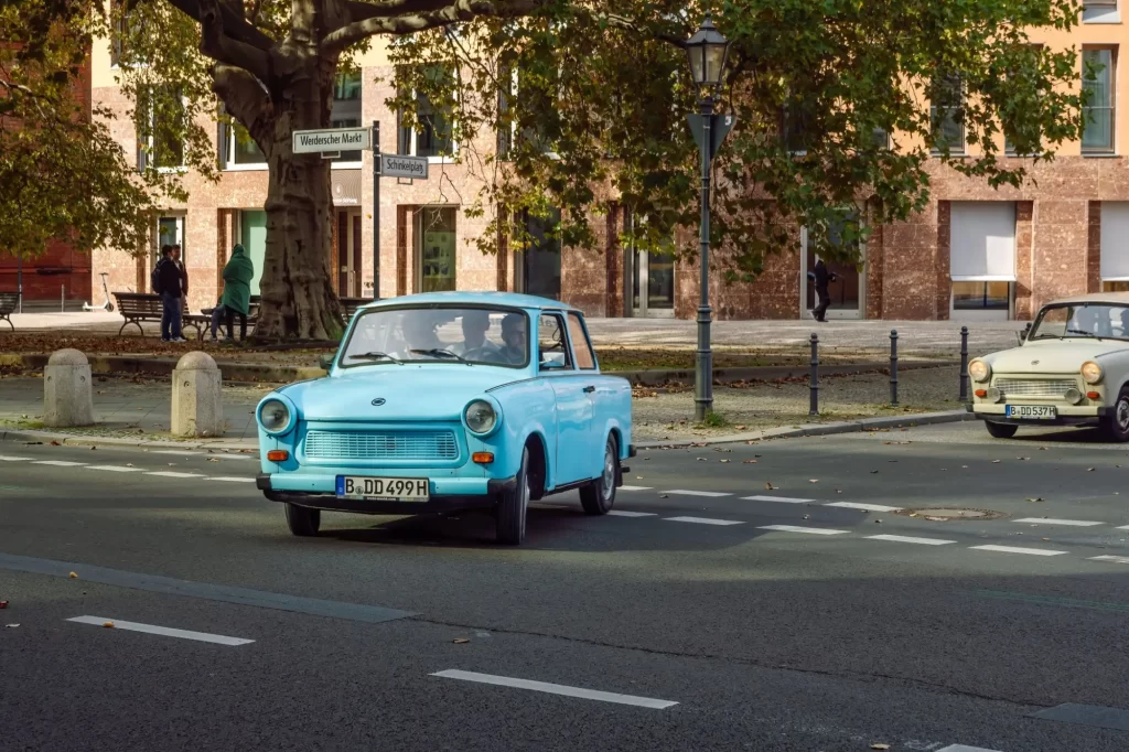 eco druhá - Techsvět Trabant projíždějící městskou ulicí lemovanou stromy a budovami.