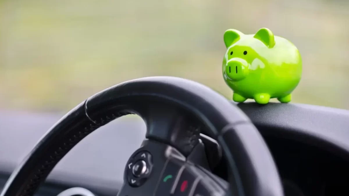 Interiér auta s detailem na volant a malou zelenou pokladničku ve tvaru prasátka, která stojí na palubní desce.
