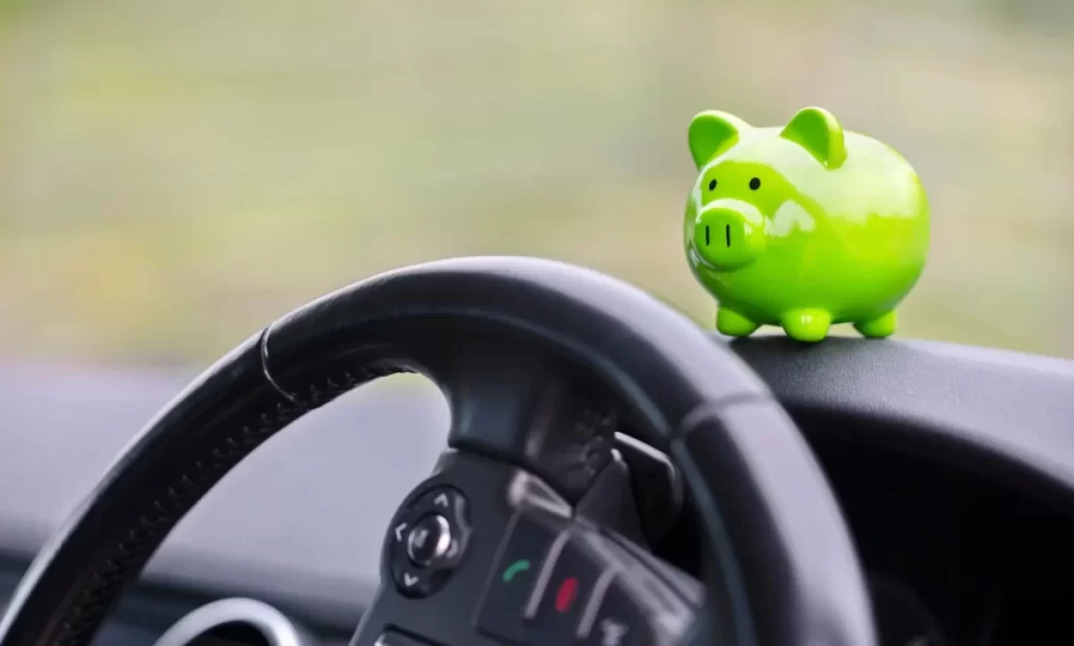 Interiér auta s detailem na volant a malou zelenou pokladničku ve tvaru prasátka, která stojí na palubní desce.