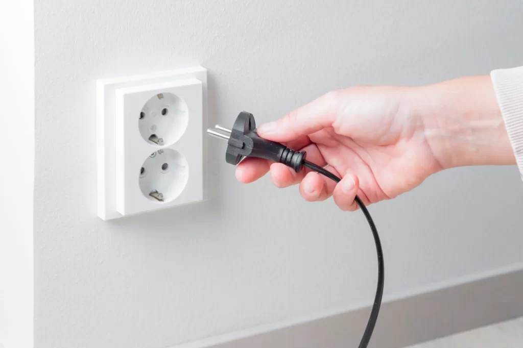 ele2 - Techsvět Ruka držící černý elektrický kabel s vidlicí, který se chystá zapojit do bílé dvojzásuvky umístěné na světlé zdi.