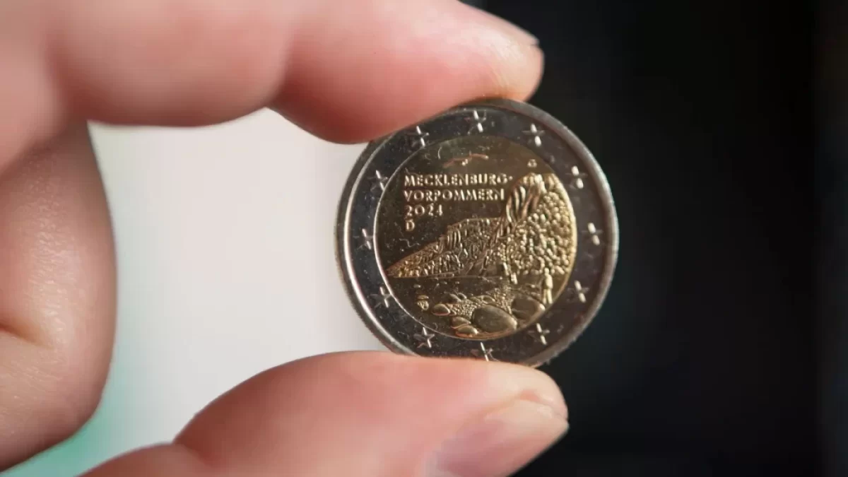 Detailní záběr na dvoueurovou minci, kterou muž drží mezi palcem a ukazováčkem.