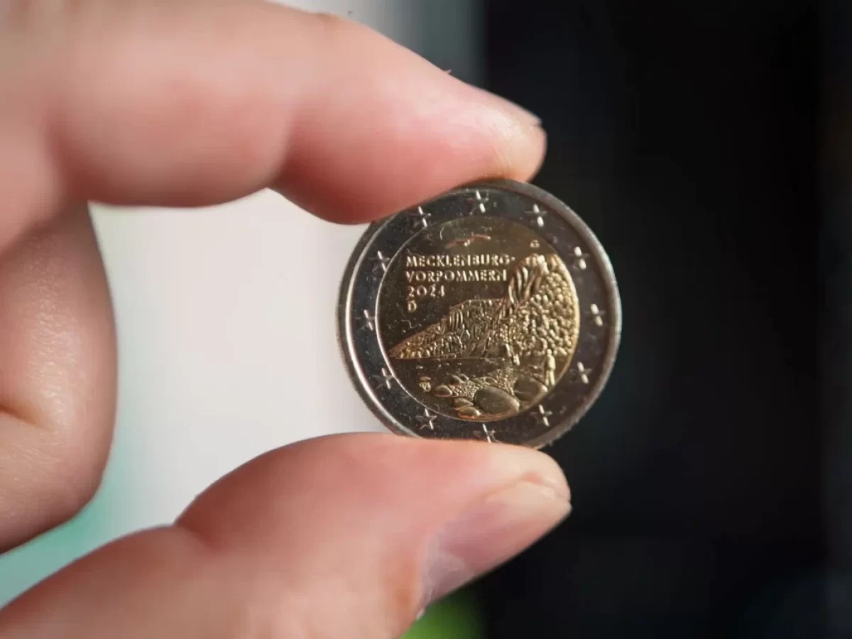Detailní záběr na dvoueurovou minci, kterou muž drží mezi palcem a ukazováčkem.