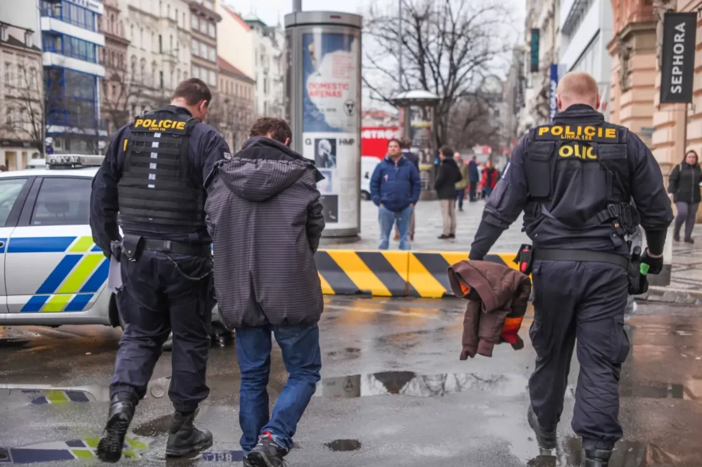 hacker3 - Techsvět Muž v civilním oblečení je veden k policejnímu vozu, zatímco okolo procházejí lidé.
