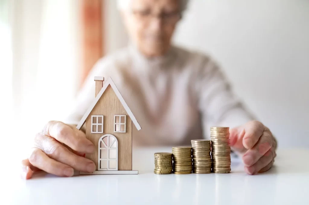 investice2 - Techsvět Starší žena před sebou drží dřevěný model domu a několik sloupců mincí.