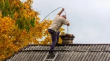 Muž stojí na střeše starší budovy a provádí čištění komína pomocí komínové štětky na laně.