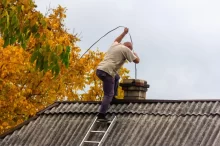 Muž stojí na střeše starší budovy a provádí čištění komína pomocí komínové štětky na laně.