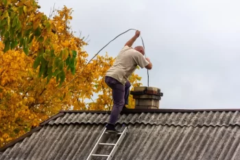 Muž stojí na střeše starší budovy a provádí čištění komína pomocí komínové štětky na laně.