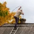 Muž stojí na střeše starší budovy a provádí čištění komína pomocí komínové štětky na laně.