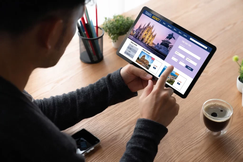 letenky2 - Techsvět Muž sedící u stolu, který prohlíží nabídky cestovní na tabletu.