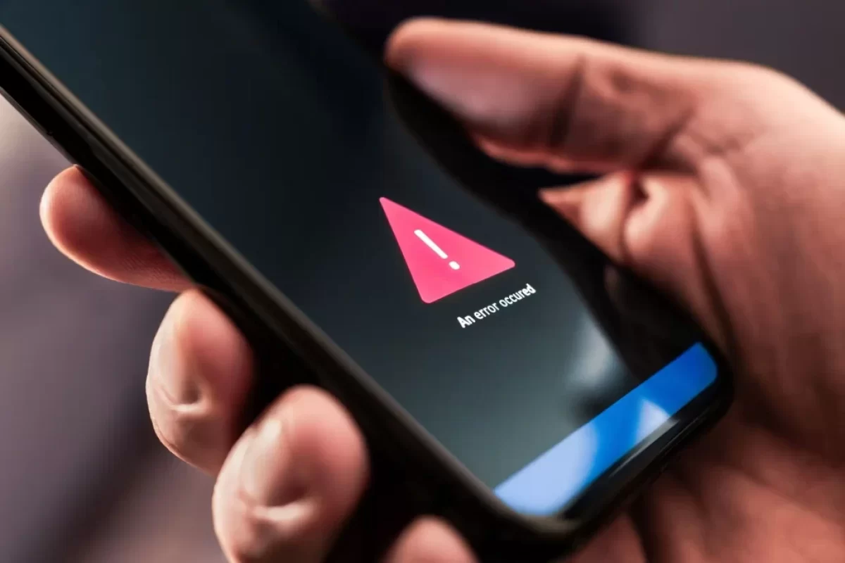 Detailní záběr na ruku držící chytrý telefon. Na displeji telefonu je zobrazený výstražný trojúhelník s vykřičníkem.