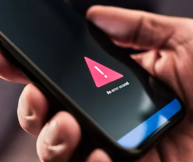 Detailní záběr na ruku držící chytrý telefon. Na displeji telefonu je zobrazený výstražný trojúhelník s vykřičníkem.