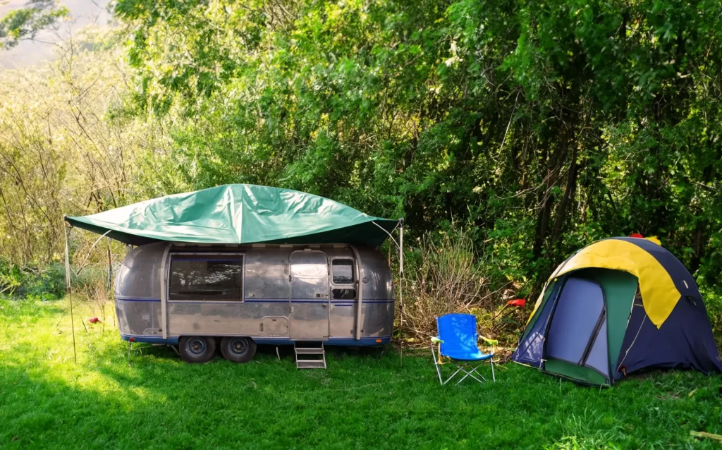 park3 - Techsvět Karavan se zelenou plachtou stojí zaparkovaný na travnaté ploše u lesa, vedle něj je postavený malý stan a rozložená modrá kempingová židle.