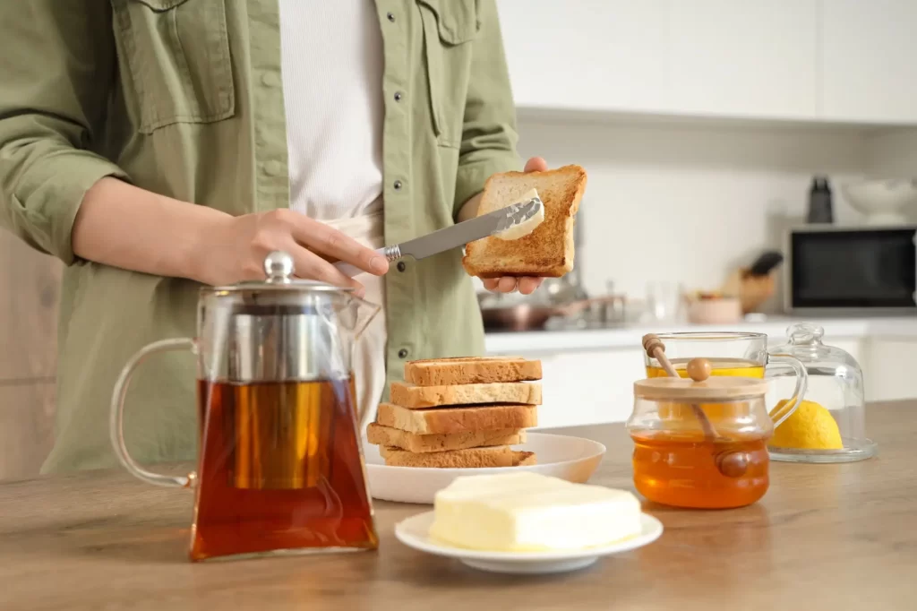 pek3 - Techsvět Žena natírá máslo na opečený toast, přičemž na stole před ní je připravený čaj v elegantní skleněné konvici, miska s máslem, sklenice medu a plátky citronu pod skleněným poklopem.