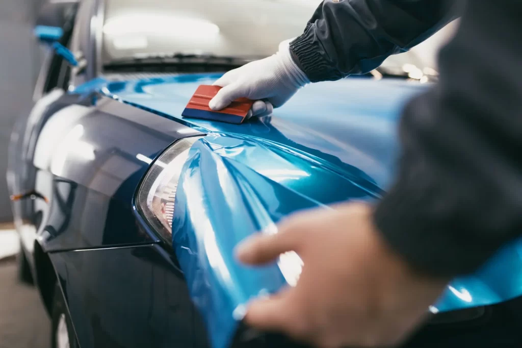 polep3 - Techsvět Technik pomocí stěrky pečlivě vyhlazuje modrou lesklou fólii na kapotu vozu.