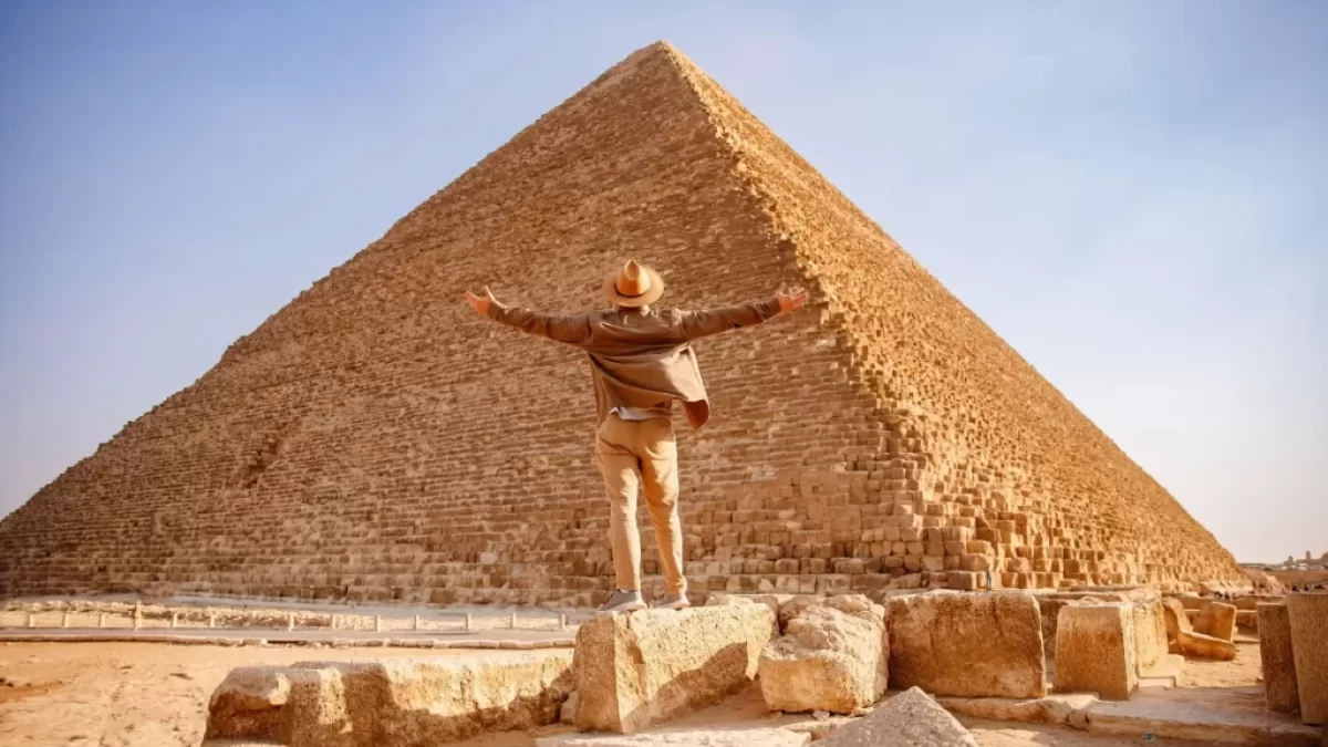 Muž s rozpaženýma rukama před monumentální Cheopsovou pyramidou v Gíze.
