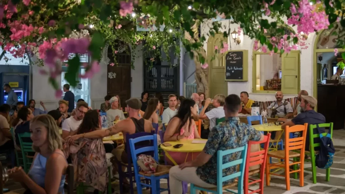 Rušná řecká taverna plná lidí, kteří si užívají večerní atmosféru pod rozkvetlými větvemi.
