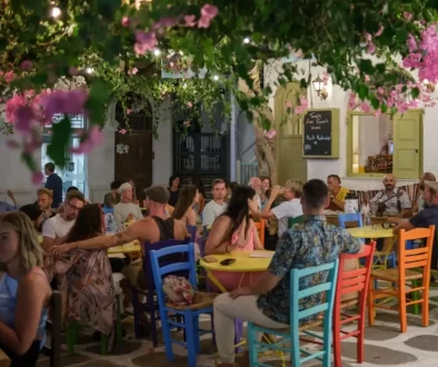 Rušná řecká taverna plná lidí, kteří si užívají večerní atmosféru pod rozkvetlými větvemi.