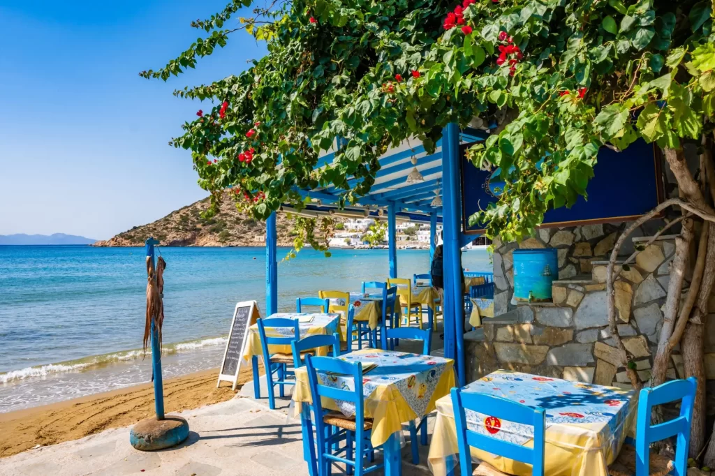 řecko2 - Techsvět Klidná řecká taverna přímo u moře, s typickými modrými židlemi, květinovou pergolou a výhledem na zátoku s písčitou pláží.