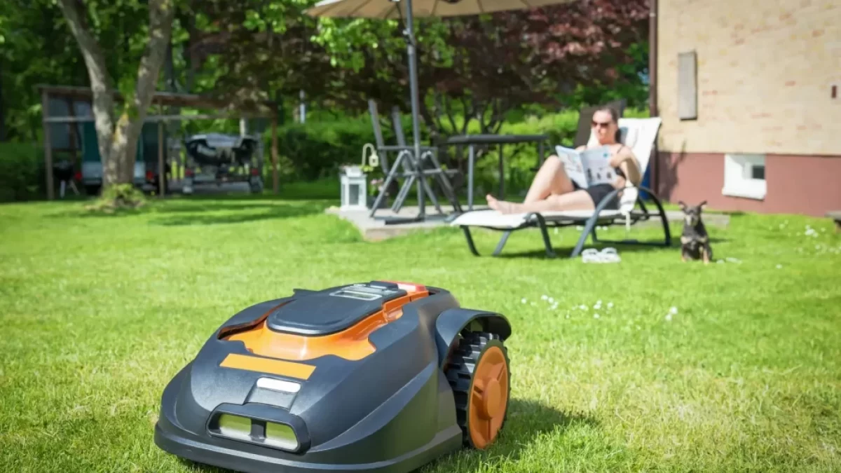 Robotická sekačka, která právě seká trávník na zahradě. V pozadí odpočívá žena na lehátku pod slunečníkem, čte noviny a vedle ní sedí malý pes.