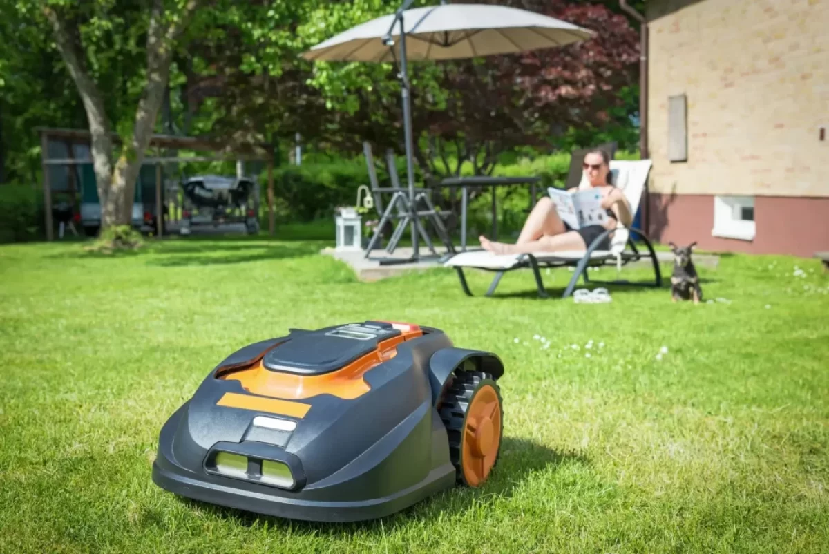 Robotická sekačka, která právě seká trávník na zahradě. V pozadí odpočívá žena na lehátku pod slunečníkem, čte noviny a vedle ní sedí malý pes.