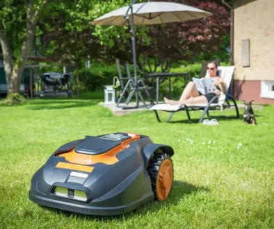 Robotická sekačka, která právě seká trávník na zahradě. V pozadí odpočívá žena na lehátku pod slunečníkem, čte noviny a vedle ní sedí malý pes.