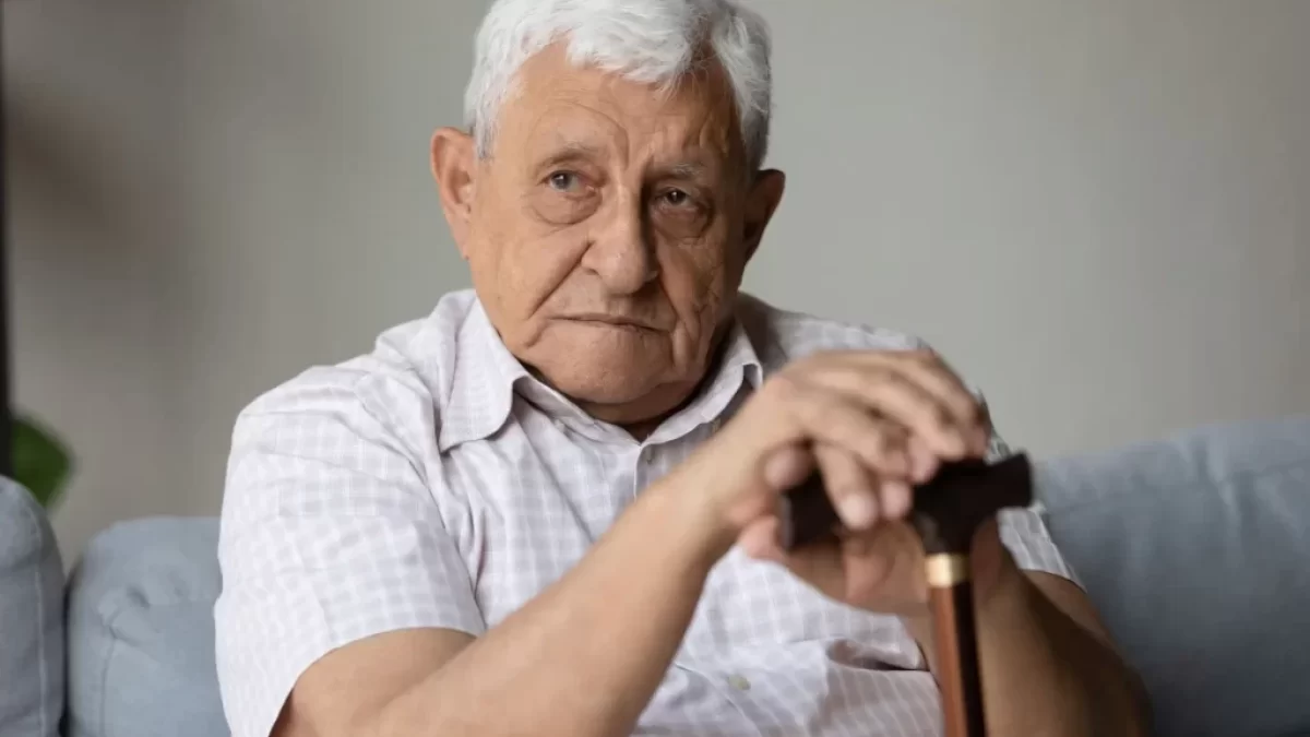 Senior v bílé košili s hůlkou sedí na gauči.
