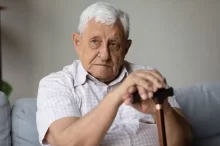 Senior v bílé košili s hůlkou sedí na gauči.