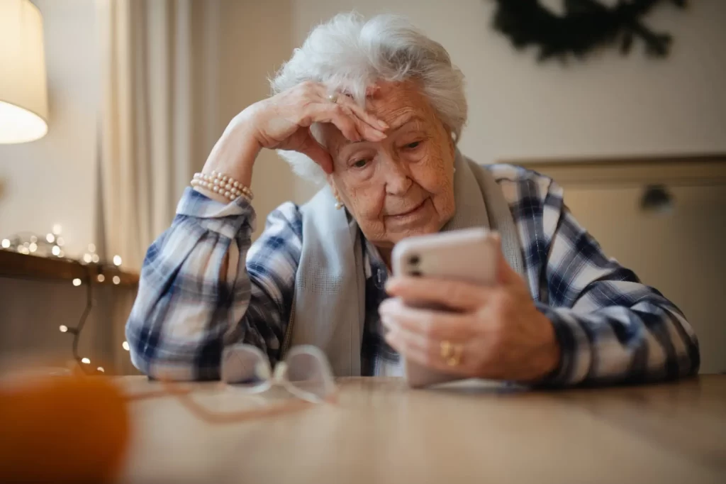 sen2 - Techsvět Starší žena sedí u stolu, drží mobilní telefon a vypadá zamyšleně nebo znepokojeně.